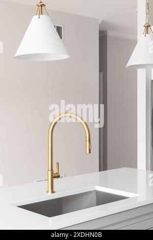 Ein Küchenwaschbecken mit goldenem Wasserhahn, weißen Schränken, Marmorarbeitsfläche und weißen und goldenen Pendelleuchten hängen über der Insel. Stockfoto