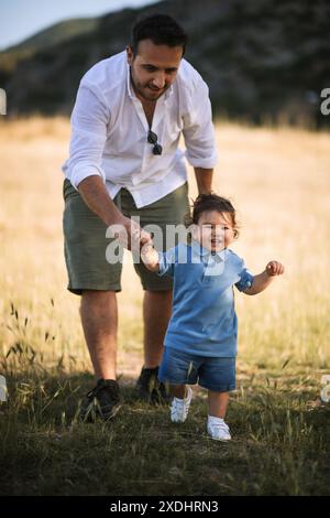 Ein Vater, der die Hand seines einjährigen Sohnes hält, während der Junge auf dem Gras läuft und beide einen freudigen Moment im Freien genießen Stockfoto