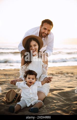 Ein kleiner Junge mit weißem Hemd und Hut sitzt auf dem Sand, seine Mutter in einem weißen Kleid hinter ihm und sein Vater lächelt hinter ihr. Sie genießen eine Schönheit Stockfoto