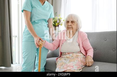Die Krankenschwester übergibt den Stock an eine ältere Frau und hilft ihr, sich in Einem Pflegeheim im Raum zu bewegen Stockfoto