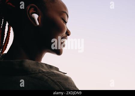 Glückliche junge Afrikanerin in ihren 20ern trägt schnurlose Ohrhörer und lächelt mit geschlossenen Augen. Sie hört gern Musik auf weißem Hintergrund Stockfoto