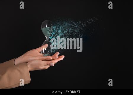 Geschäftsfrau mit auflösender Sanduhr auf schwarzem Hintergrund, Nahaufnahme. Die Zeit läuft ab Stockfoto