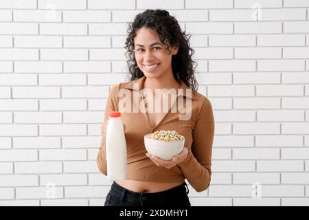 Glückliche junge Frau, die eine Schüssel mit leckeren Müsliringen und Milch in der Nähe einer hellen Ziegelwand hält Stockfoto