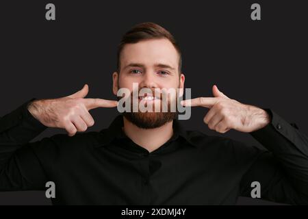 Junger Mann, der auf seinen Bart zeigt, auf dunklem Hintergrund Stockfoto