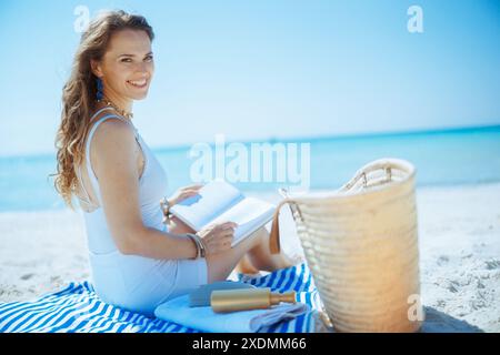 Lächelnde, elegante Frau mittleren Alters am Meer mit Strohbeutel, Buch und gestreiftem Handtuch. Stockfoto