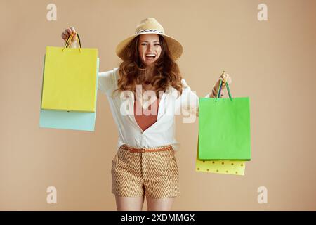 Strandurlaub. Lächelnde, elegante Frau mittleren Alters in weißer Bluse und Shorts isoliert auf Beige mit Einkaufstasche und Sommerhut. Stockfoto