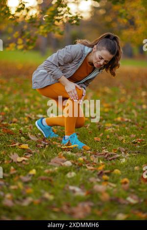 Hallo Herbst. Unglücklich fit Frau mittleren Alters in Fitnesskleidung im Park mit Beinschmerzen. Stockfoto