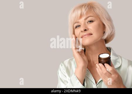 Schöne reife Frau im Pyjama mit Glas Gesichtscreme auf grauem Hintergrund Stockfoto