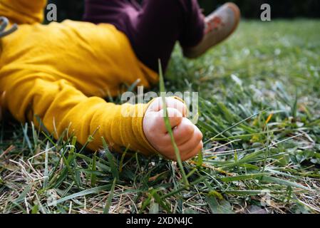 Ein Kind, das einen gelben Pullover trägt, fällt auf das Gras, wobei es sich auf die Hand des Kindes konzentriert und einen Moment der Verspieltheit festnimmt Stockfoto