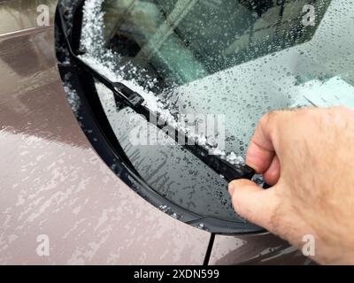 Eine Hand, die einen Scheibenwischer an einem Auto hochhebt und Hagel und Regentropfen auf der Windschutzscheibe zeigt, was auf einen Hagelsturm hindeutet, mit dem Ex Stockfoto