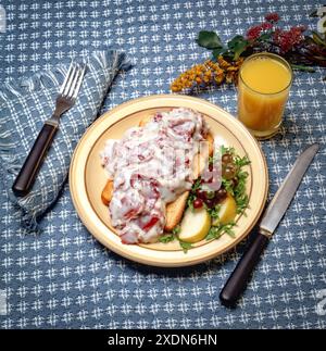 Frühstücksteller mit cremigem Rindfleisch auf Toast mit cremiger Soße, serviert mit Trauben, Birnenscheiben und Orangensaft auf rustikalem blauem Tisch. Stockfoto