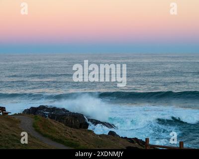 Schichten von herrlichem rosafarbenem, hellorangenem und blauem Pastellhimmel spiegeln sich in Wolken im Osten über dem Pazifik, mit stürzenden Wellen, Australien Stockfoto