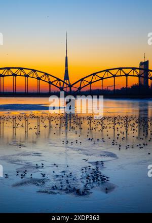 Eine wunderschöne Landschaft bei Sonnenaufgang mit eiserner Brücke über den gefrorenen Fluss Daugava in der lettischen Hauptstadt Riga. Winterlandschaft Nordeuropas. Stockfoto