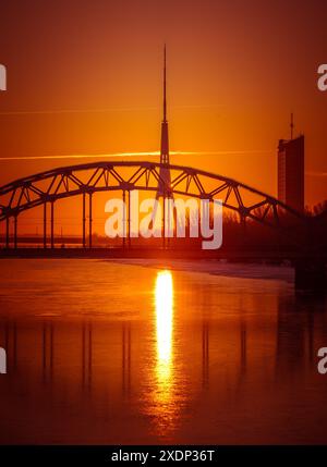 Eine wunderschöne Landschaft bei Sonnenaufgang mit eiserner Brücke über den gefrorenen Fluss Daugava in der lettischen Hauptstadt Riga. Winterlandschaft Nordeuropas. Stockfoto