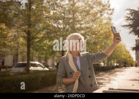 Eine Frau fotografiert sich mit ihrem Handy. Sie trägt eine graue Jacke und einen Schal. Die Szene spielt in einer Stadt mit Bäumen und Autos Stockfoto