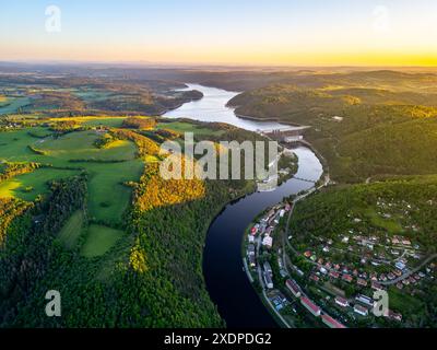 Ein Luftbild des Orlik Reservoir in Tschechien bei Sonnenuntergang zeigt einen gewundenen Fluss, Hügel und eine malerische Stadt im Tal mit leuchtendem blauem und goldenem Himmel. Stockfoto