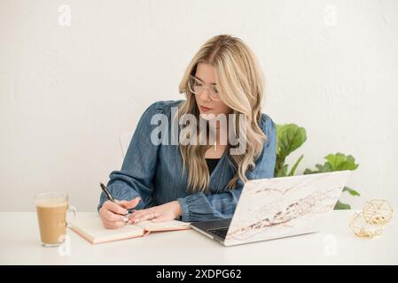 Frau, die in Notizbuch mit Kaffee und Laptop des Schreibtisches schreibt Stockfoto