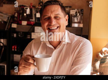 Mann mit weißem Hemd lächelt, während er eine weiße Kaffeetasse hält Stockfoto