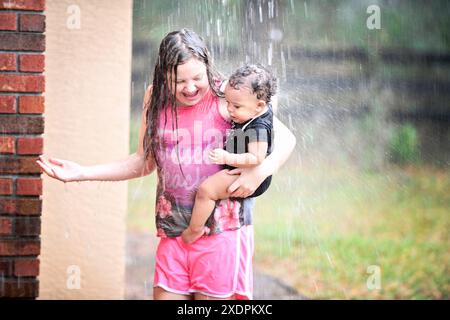 Mädchen und Baby spielen im Regen, beide lächeln und genießen. Stockfoto