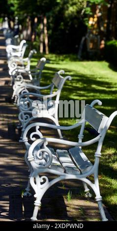 Weiße Bänke in einem Stadtpark, eingebettet im Schatten von Bäumen an einem sonnigen Tag, bieten einen ruhigen und ruhigen Ort für Ruhe und Entspannung Stockfoto