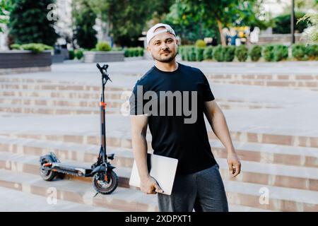 Schöner Mann hält Laptop in der Hand und steht in der Nähe eines Elektrorollers. Aktiver Lebensstil, Rucksacktourist. Ökologie und saubere ökologische Modus der tr Stockfoto