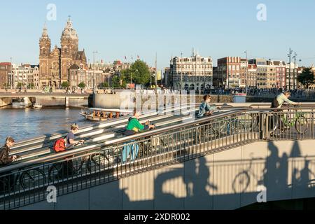 Radfahrer schieben ihre Fahrräder vom unterirdischen Fahrradparkplatz in der Centraal Station, mit dem Kanal und der Basilika St. Nikolaus im Hintergrund, in Amsterdam, Niederlande. Stockfoto