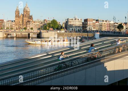 Radfahrer schieben ihre Fahrräder vom unterirdischen Fahrradparkplatz in der Centraal Station, mit dem Kanal und der Basilika St. Nikolaus im Hintergrund, in Amsterdam, Niederlande. Stockfoto