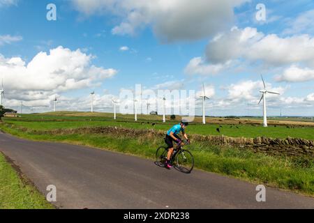 Erfahrener Radfahrer, der an einem sonnigen Frühlingstag auf einer Landstraße in South Yorkshire entlang einer Trockenmauer fährt, mit Windturbinen und Wolken dahinter Stockfoto