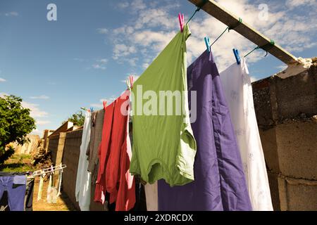 Farbenfrohe Kleidung hängt an einigen Seilen mit Kleiderschalen, im Innenhof des Dorfhauses, um an einem sonnigen Tag an der Luft zu trocknen, mit einigen Wolken im Blau Stockfoto