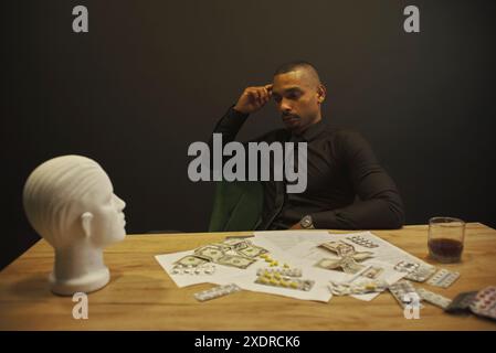 Ein nachdenklicher Afroamerikaner sitzt an einem Tisch und schaut auf den Kopf einer Schaufensterpuppe. Auf dem Tisch sind Geld und Pillen verstreut und Whiskey in einem gl Stockfoto