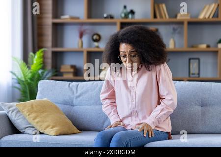 Frau auf der Couch, die ängstlich und beunruhigt erscheint und wahrscheinlich Panikattacken erlitten hat. Modernes Wohnzimmer mit Bücherregal Hintergrund. Sie trägt ein rosafarbenes Hemd mit ausdrucksstarker Gesichtsreaktion. Stockfoto