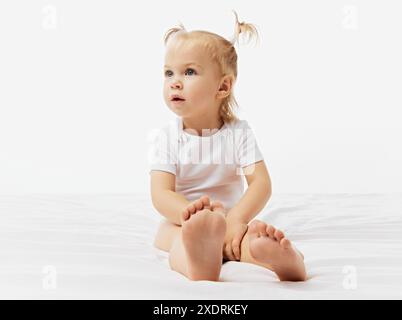 Kurioses Kind, kleines Mädchen mit blonden Zöpfen sitzt auf weißer Oberfläche, gekleidet in Einteiler vor weißem Studiohintergrund. Stockfoto