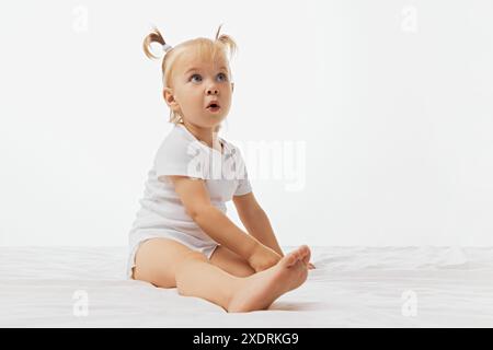 Porträt eines Kleinkindes mit blonden Zöpfen, gekleideter Einteiler sitzt auf weißer Oberfläche mit einem Ausdruck des Staunens vor weißem Studiohintergrund. Stockfoto