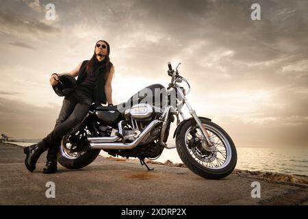 Ein Mann im Alter von 30 Jahren in Metallkleidung lehnt sich an sein Motorrad mit einem malerischen Blick auf das Meer bei Sonnenuntergang. Stockfoto