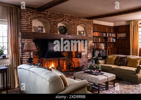 Feuer mit Ornamenten auf einem Kaminmantel aus Ziegelstein mit Bücherregalen und Sofas im Tudor Farmhouse aus dem 16. Jahrhundert, Hertfordshire, England, Großbritannien. Stockfoto