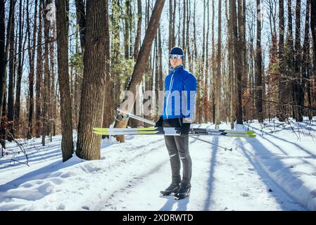 Langlaufläufer in blauer Jacke stehen im verschneiten Wald und halten Ski bereit für ein Outdoor-Abenteuer. Männlicher Athlet ging zum Tagestraining im Park auf der Skipiste. Stockfoto