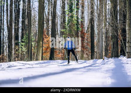 Skilanglauf in blauer Jacke gleitet am sonnigen Wintertag durch verschneite Wälder und genießt Outdoor-Aktivitäten. Man Athlet trainiert auf Skiern, indem man auf der vorbereiteten Strecke Schlittschuhlaufen kann. Stockfoto