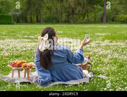 Glückliche Frau genießt das Leben. Verbringt Zeit in der Natur. Eine Dame sitzt auf dem Gras in einem Park, mit einem Picknicktisch mit Essen in der Nähe. Rückansicht. Es gibt einen Stockfoto