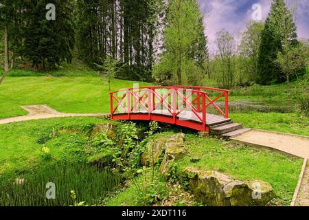 Ein Park mit üppigem Laub wird durch eine rote Holzbrücke hervorgehoben, die elegant einen strem in Grewelthorrpe, Ripon, Großbritannien, überspannt. Stockfoto