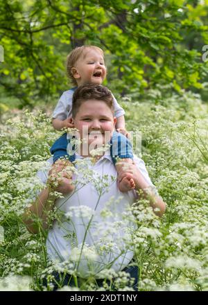 Porträt eines älteren und eines kleinen Bruders im dicken, grünen Gras. Ein glücklicher Junge hält ein fröhlich lachendes kleines Baby auf den Schultern mit welligen, blonden Bäumen Stockfoto