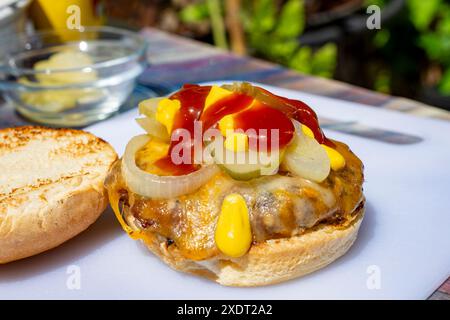 Direkt vom Grill, Burger Patty garniert mit Cojakkäse, gegrillten Zwiebeln, Dill-Gurken, Ketchup und Senf auf einem gerösteten Brötchen. Stockfoto