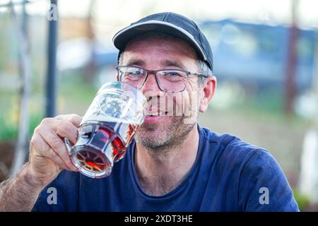 Ein älterer Mann hält ein 500-ml-Glas, das speziell entwickelt wurde, um das erfrischende Bier im Sommer zu genießen. Er hat eine Mütze auf dem Kopf und ein blaues T Stockfoto
