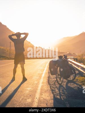 Der Radsportler steht selbstbewusst und entschlossen Hand an Kopf und blickt mit dem Fahrrad im Stehen in die Berge. Reisen Sie durch Georgia Kazbegi na Stockfoto