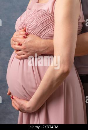 Schwangere Frau in einem rosa Kleid auf blauem Hintergrund. Der Ehemann ist zurück. Hände auf dem Bauch halten. Keine Fläche. Nahaufnahme. Stockfoto