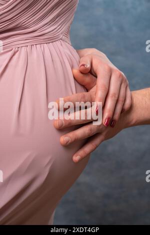 Auf dem Bauch einer schwangeren Frau in rosa Kleid liegt die Hand eines Musikers. Nahaufnahme. Blauer Hintergrund Stockfoto