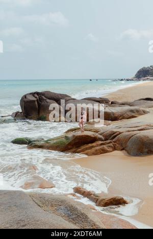 Eine Frau steht an einem felsigen Strand in Thailand, mit einem klaren blauen Ozean im Hintergrund. Die Flut kommt, und die Wellen schlagen sanft auf die Stockfoto