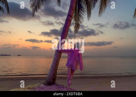 Eine Frau lehnt sich an eine Palme am Strand, während die Sonne über dem Meer untergeht. Der Himmel ist leuchtend orange und rosa, und das Wasser ist ruhig, hellblau Stockfoto