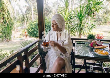 Eine Frau mit weißem Bademantel sitzt auf einer Veranda und genießt eine Tasse Tee. Sie blickt auf das üppige tropische Grün. Die Veranda hat eine hölzerne Schiene Stockfoto