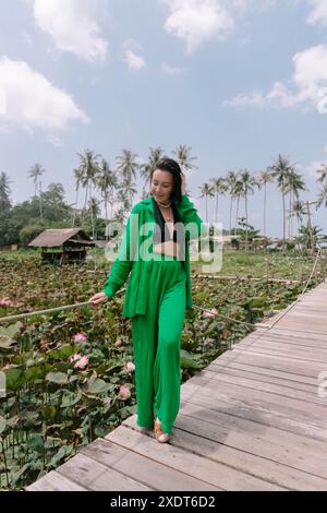 Schöne Frau in grünem Outfit spaziert entlang einer Holzbrücke über einen Seerosenteich, umgeben von Palmen und üppiger Vegetation. Stockfoto