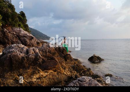 Eine Frau sitzt an einem felsigen Ufer bei Sonnenuntergang und blickt auf das ruhige Meer. Üppiges Grün und eine ferne Bergkette bieten eine malerische Kulisse. Stockfoto